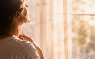 Mulher diante de uma janela iluminada respirando fundo, mão no peito — culpa sendo liberada