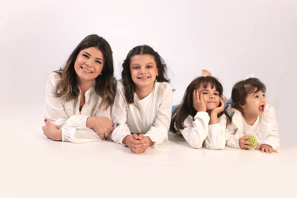Samara deitada no chão com as três filhas, todas de branco, rindo